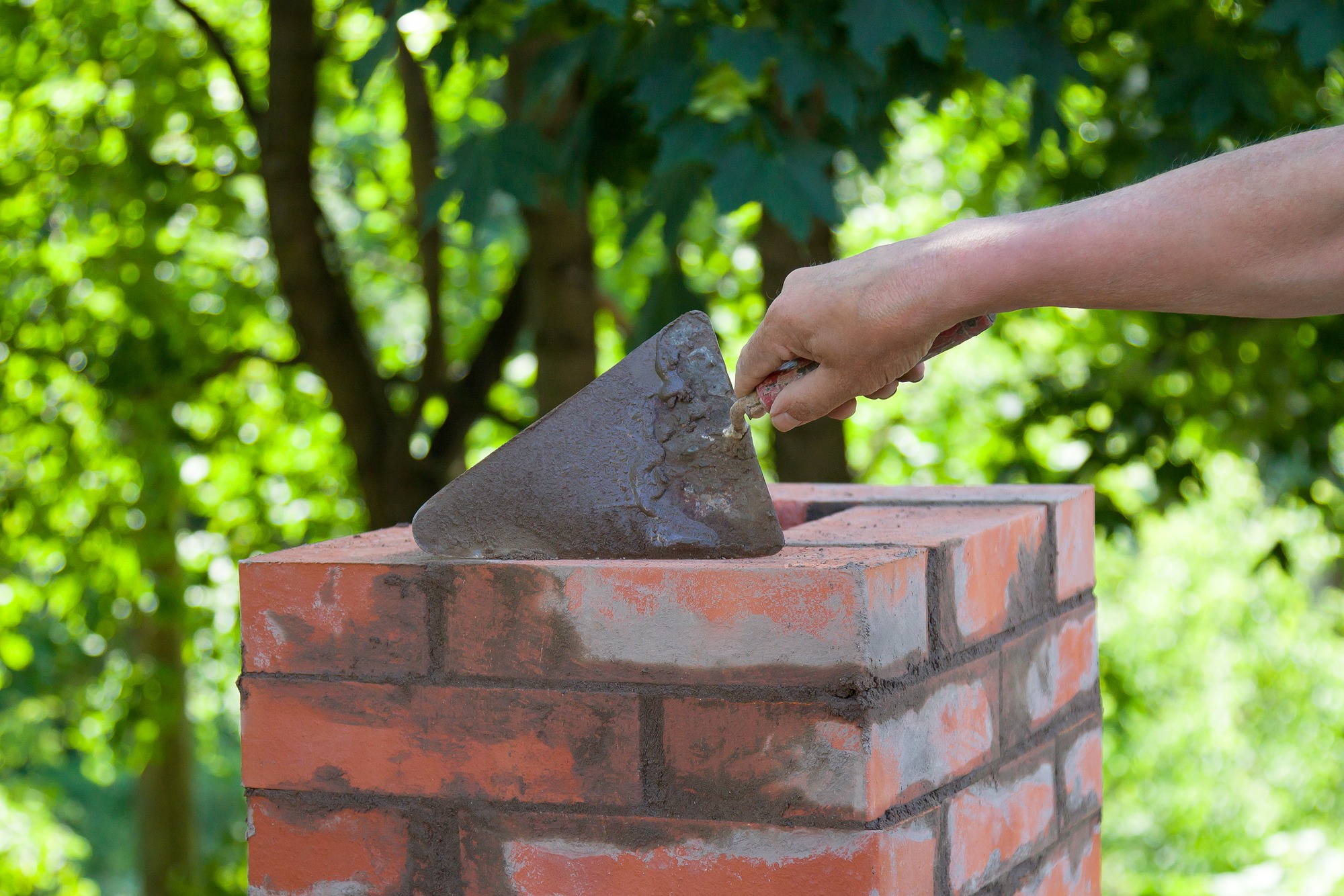 A person's hand holds a trowel as they apply mortar to the top of a brick wall in a lush outdoor setting, showcasing masonry work in progress.