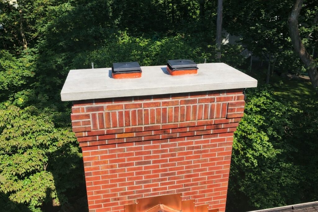 Chimney with a concrete cap topped by two black flue caps, surrounded by lush green trees. Highlights a well-maintained rooftop feature.