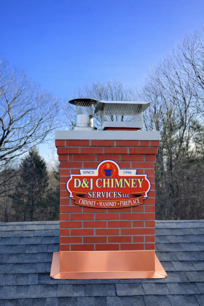 D&J Chimney Services sign on a brick chimney, featuring a metal cap, atop a roof under a clear blue sky. Illustrates chimney maintenance services.