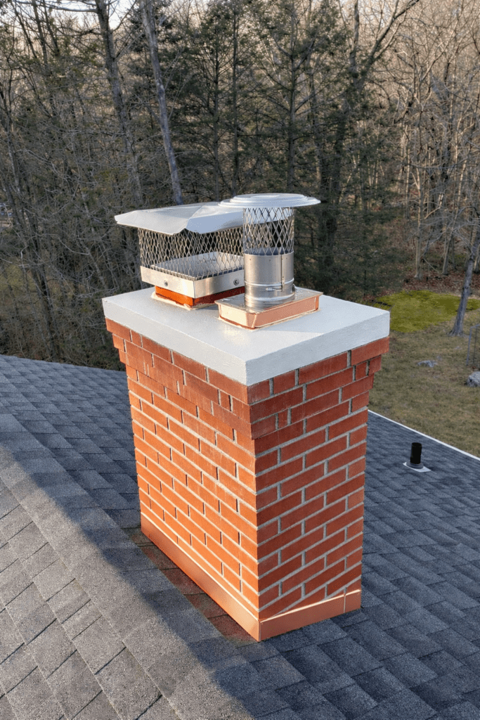 A red brick chimney with a shiny metal cap on a dark shingled roof. Surrounding trees appear bare, suggesting a winter setting.