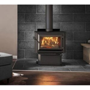 Cozy room with a wood-burning stove against a dark stone wall. Logs are stacked nearby. A gray armchair and soft lighting create a warm, inviting atmosphere.